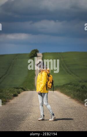 Woman hiking on road and looking at cloudy sky. Rain is coming. Backpacker wearing hat and yellow waterproof jacket Stock Photo