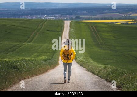 Long journey. Hiking woman walking on empty road. Hiker with hat and backpack wearing yellow waterproof jacket. Travel concept. Getting away from it a Stock Photo