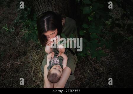 A little baby lies on the lap of her mother, who in turn kisses her daughter's sweet feet. Love, day and motherhood concept. Walking a happy family in Stock Photo