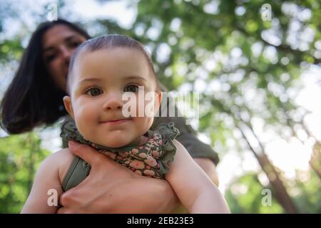 Young brown-haired mother shakes her baby in her arms in the park among the trees, green grass. Woman holds little girl in her arms, plays, hugs child