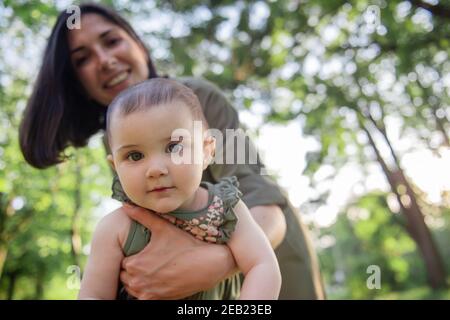 Young brown-haired mother shakes her baby in her arms in the park among the trees, green grass. Woman holds little girl in her arms, plays, hugs child Stock Photo