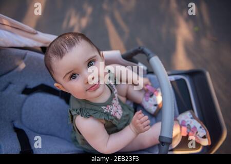 A cute little baby girl with green eyes in a dress sits in a stroller among a green park. Close-up portrait of a child looking into the camera. Copy s Stock Photo