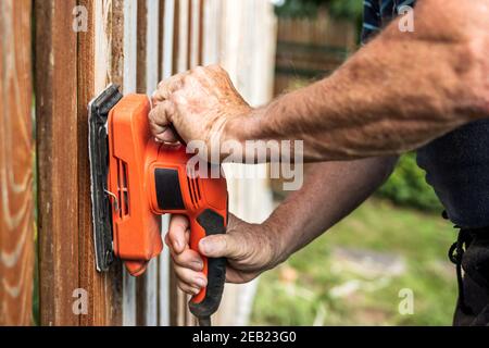 Sanding wood. Close-up sander in male hands. Repairing wooden fence at backyard Stock Photo