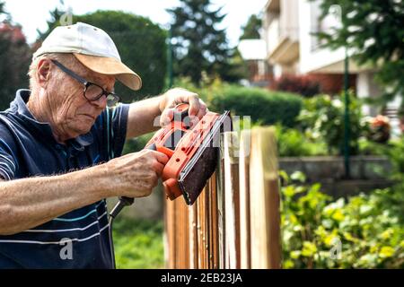 Sander in hands. Senior man sanding wooden fence in garden. Old carpenter using electric work tool Stock Photo
