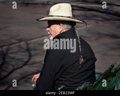 An Hispanic man with braided hair (Ramon Jose Lopez, a well-known artist) sits on a bench in the historic Plaza in Santa Fe, New Mexico. Stock Photo