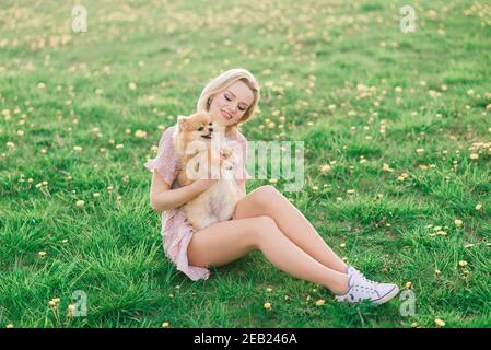 Portrait of a young glamorous female wearing pink dress holding her cute pomeranian spitz on hands. . Friendship between dogs and humans. Stock Photo