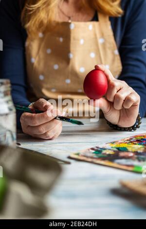 Woman wearing apron and coloring Easter eggs for traditional religious holiday. Red egg in female hand. Artist is ready for painting homemade decorati Stock Photo