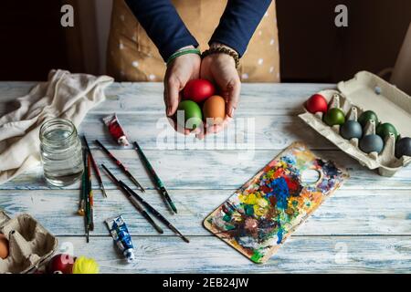 Ready to paint Easter egg. Woman holding colorful eggs in her hands. Decorating for traditional religious celebration. White wooden table with paintbr Stock Photo