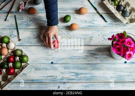 Preparation for painting Easter eggs. Female hand, paintbrush, colorful eggs and pink primrose flower on white wooden table. Flat lay Stock Photo