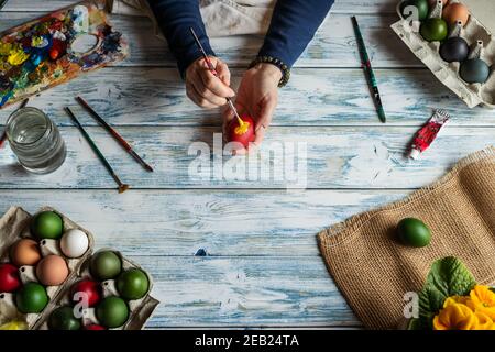 Hands painting Easter eggs on white wooden table, top view. Woman coloring egg for traditional religious celebration. Artist using colors, paintbrush Stock Photo