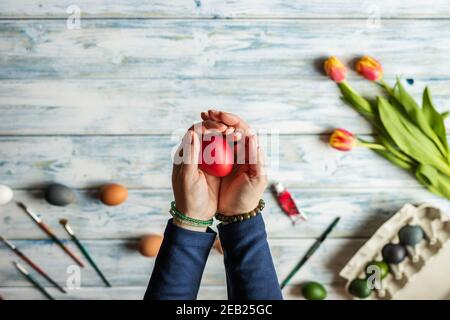 Woman holding coloring easter egg in her hands. White wooden table with tulip flower. Decorating for Easter holiday. Top view Stock Photo