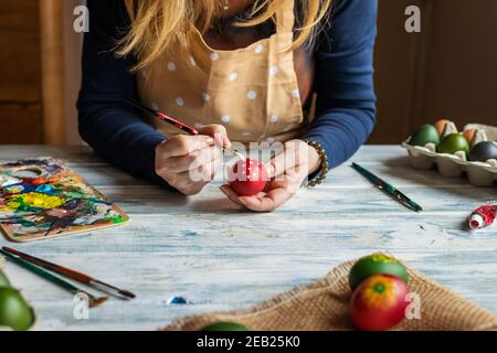 Woman painting egg and preparing for Easter holiday celebration. Paintbrush in female hand and palette on white wooden table. Decorating at home Stock Photo