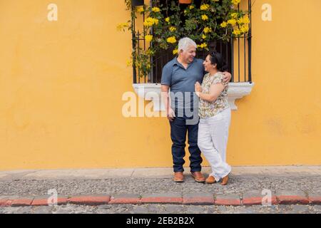 Adult couple embracing looking at each other on a yellow wall with flowers in Antigua Guatemala- Senior couple in love on vacation in colonial city Stock Photo