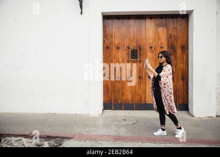 Full portrait of Hispanic woman taking a picture with her phone in colonial city-tourist taking pictures while walking in Antigua Guatemala Stock Photo