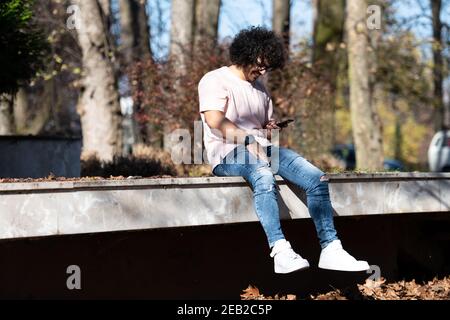 Portrait of Handsome Young Afro Man Talking Touching Phone or Using Smartphone for the Call and Sitting Outside During Autumn Stock Photo