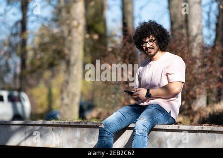 Portrait of Handsome Young Afro Man Talking Touching Phone or Using Smartphone for the Call and Sitting Outside During Autumn Stock Photo