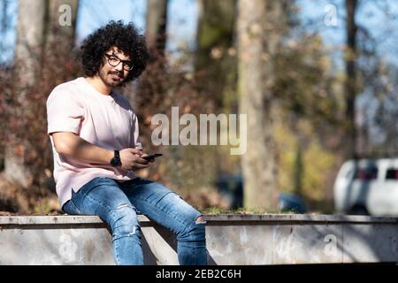 Portrait of Handsome Young Afro Man Talking Touching Phone or Using Smartphone for the Call and Sitting Outside During Autumn Stock Photo