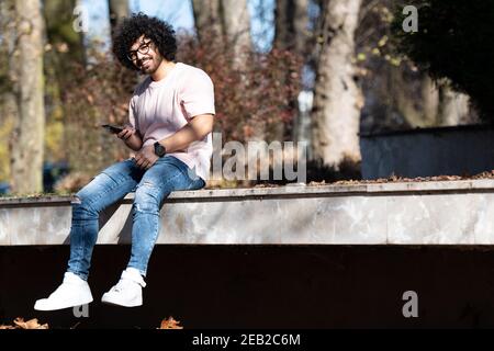Portrait of Handsome Young Afro Man Talking Touching Phone or Using Smartphone for the Call and Sitting Outside During Autumn Stock Photo