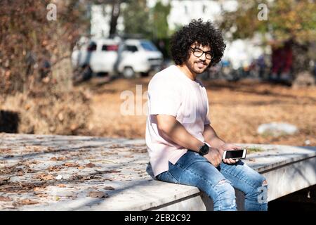Portrait of Handsome Young Afro Man Talking Touching Phone or Using Smartphone for the Call and Sitting Outside During Autumn Stock Photo