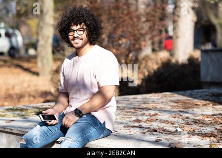 Portrait of Handsome Young Afro Man Talking Touching Phone or Using Smartphone for the Call and Sitting Outside During Autumn Stock Photo