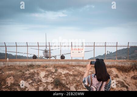 Tourist Woman is Capturing Photos The Airplane While Take off Landing on Runway Track From Outside The Airport. Asian Tourist Woman Having Fun With Ph Stock Photo