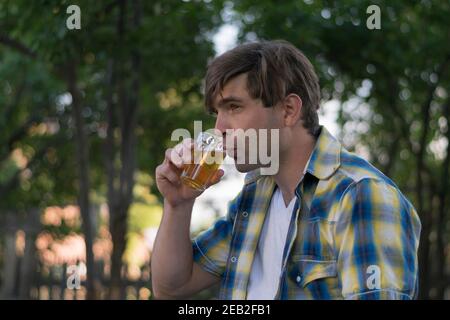 A young white man tastes beer in a glass outdoors in the shade. He is wearing a shirt of blue and yellow plaid and a white t-shirt. Stock Photo