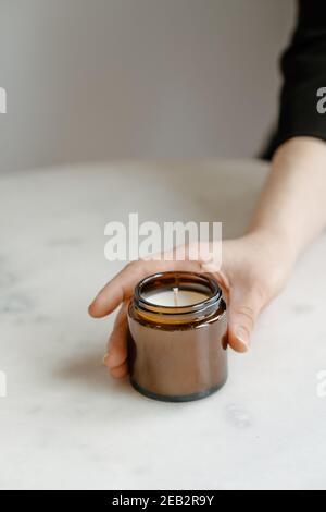 Woman's hand holding organic coconut candle in brown glass packaging on white table. Product photography concept, minimalism, copy space, still life Stock Photo