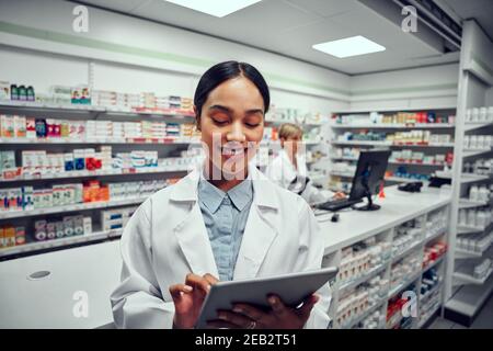 Successful young female pharmacist using digital tablet in drugstore with colleague using computer in background Stock Photo
