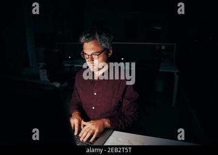 Businessman sitting in dark office room working on computer late at night Stock Photo