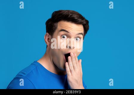 Portrait of  caucasian male model in astonished gesture with hand covering mouth in blue isolated studio background, selective focused Stock Photo