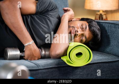 Tired young man sleeping on sofa by holding dumbbells and yoga mat - concept of healthcare, lifestyle and fitness man resting after workout in home Stock Photo