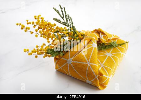 Wrapped gift by linen fabric in furoshiki eco style with yellow mimosa flower. Woman or mother day celebration concept. Stock Photo