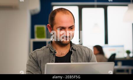 Enthusiastic business man writing a report sitting at table in modern startup business office sending mails typing on laptop, greeting colleague. Man working in financial company with diverse team. Stock Photo