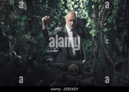 Confident corporate businessman surviving in the jungle, he is holding a machete and his briefcase Stock Photo