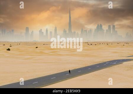 View from above, stunning aerial view of an unidentified person walking on a deserted road covered by sand dunes with the Dubai Skyline