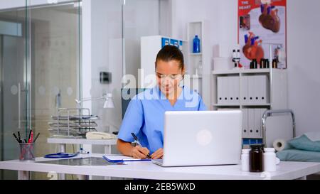 Nurse taking notes on clipboard after reading medical raports of patient from laptop. Healthcare physician in uniform, doctor assistant working in hospital checking registration, writing treatments Stock Photo