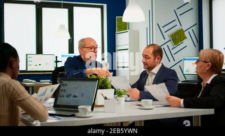 Senior businessman team leader brainstorming with colleague using paperwork showing ideas working in office. African woman using laptop in meeting broadroom, negotiations speaking with clients Stock Photo