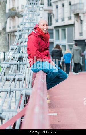 Smiling fashion hipster Hispanic man wearing a red coat and scarf sitting on a metal railing Stock Photo