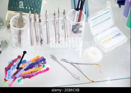 Glass table with various orthodontic instruments, bracket placement card and multicolored rubber bands. Stainless steel instruments for procedure of attaching braces on desk in dental office. Stock Photo