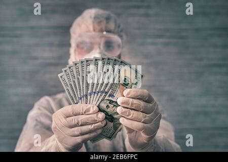 A scientist in a protective suit and medical gloves and mask holds cash in his hands. Dirt germs and virus bacteria on money. Stock Photo