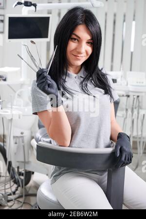 Female dentist in sterile gloves holding tools for dental treatment. Charming doctor smiling to camera while demonstrating dental instruments. Concept of dentistry, stomatology and dental medicine. Stock Photo