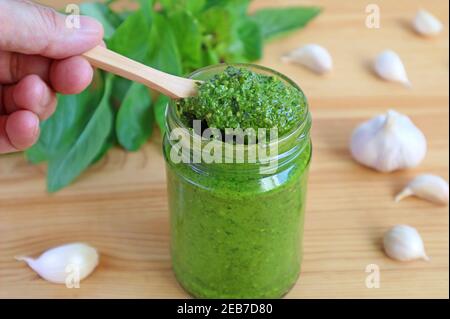 Hand Holding Wooden Spoon Scooping Fresh Basil Pesto Sauce or Pesto Alla Genovese from the Bottle Stock Photo