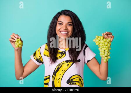 Photo of positive dark skin girl hold green organic grape branch wear pink t-shirt isolated on teal color background Stock Photo