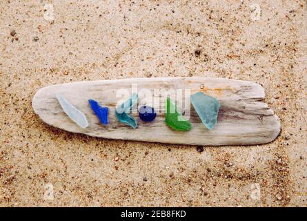 Different color blue, green, white sea glass or beach glass on driftwood in beach on sand. Glass use to be bottle pieces and now sea has tumbled. Stock Photo