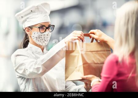 Chef wearing hat, glasses and face mask hands a paper bag with delivery food to a customer. Stock Photo