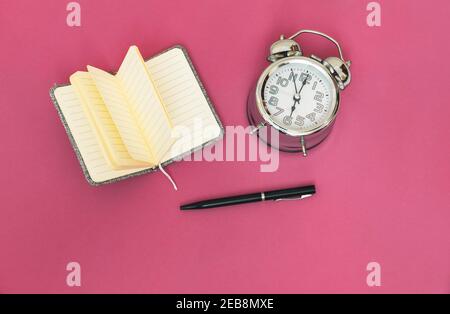 Top view of an open notebook for project notes, a black pen and an analog time concept clock for ideas on a pink background. Stock Photo