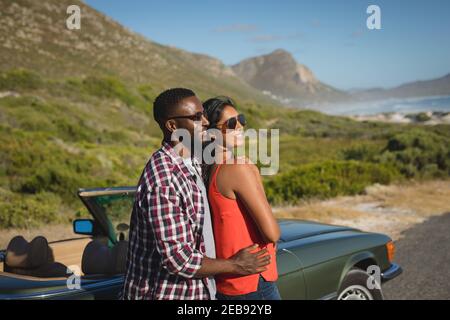 Diverse couple on sunny day standing by convertible car embracing and smiling Stock Photo
