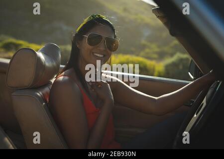 Mixed race woman driving on sunny day in convertible car holding driving wheel and smiling Stock Photo