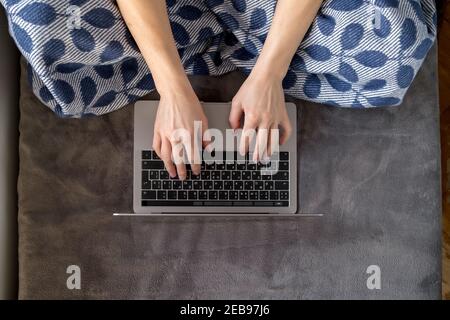 A woman wrapped in a plaid works with a laptop sitting on a soft sofa at home.  Stock Photo