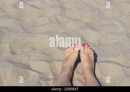 Female feet on beach sand. Woman's feet with red nails on seashore sand background in sunlight near sea. Stock Photo